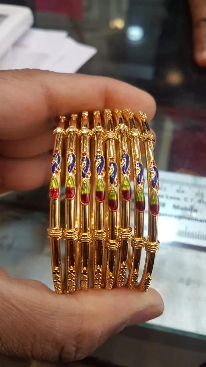 A hand holding a stack of gold bangles enamelled with peacocks in blue, green and red.