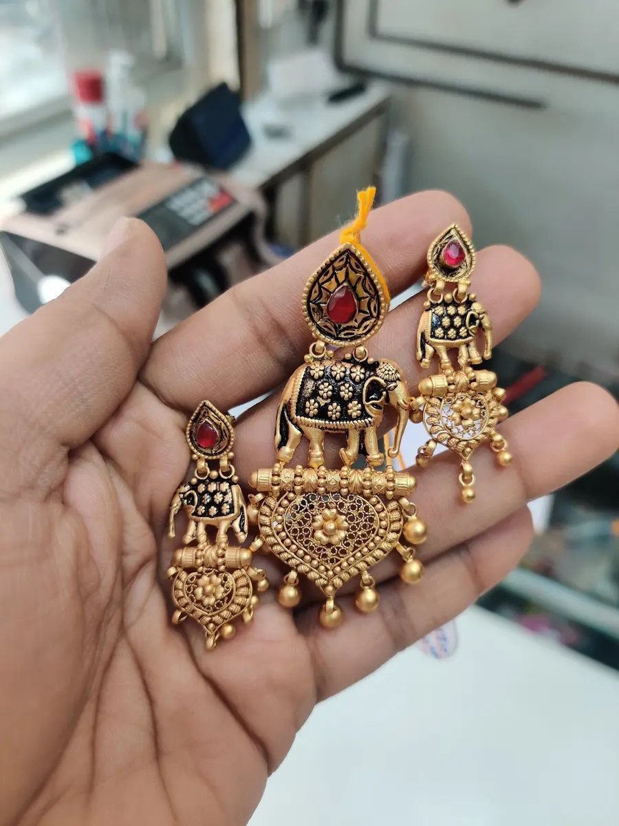 A hand holding two gold temple earrings with elephant motifs, ruby and black enamel detail.